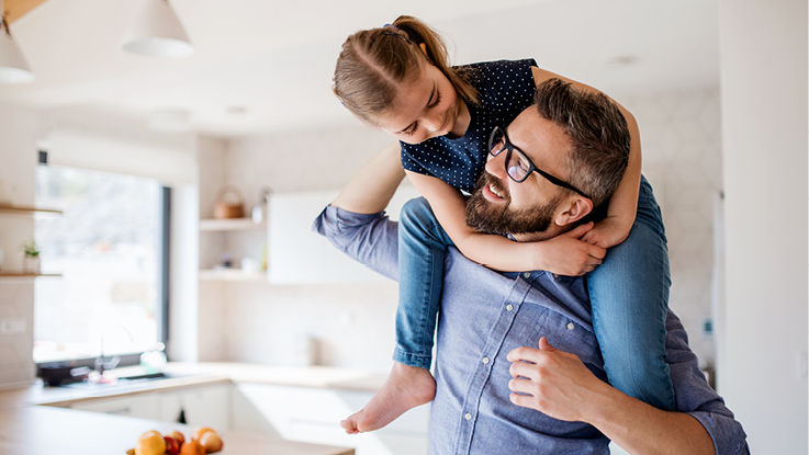 A dad balances his daughter on his shoulders and smiles in a sunny kitchen.