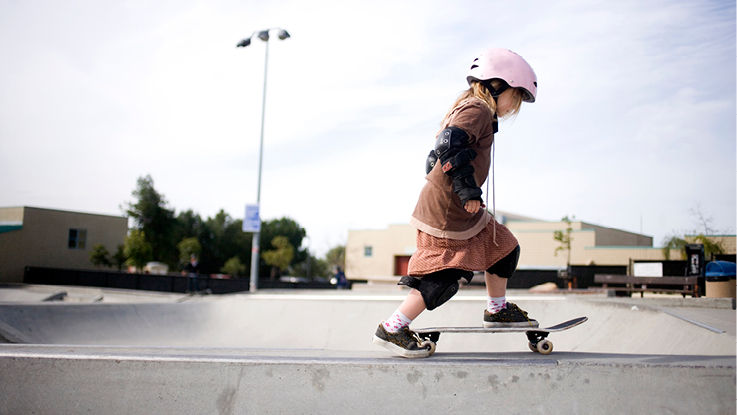 A kid wearing a pink helmet skateboards at an urban skateboard park on a cloudy day.