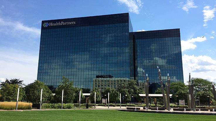 A picture of HealthPartners corporate headquarters. It’s a large, modern building covered in windows located next to a park.