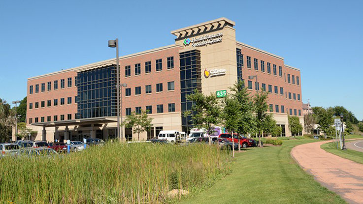 An exterior picture of the HealthPartners specialty center. The photo shows a large building with many windows, a parking lot, and a walking path that goes past the building.