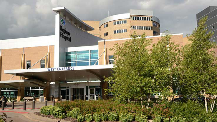 A picture of the west entrance of Regions Hospital. It’s a covered entrance surrounded by trees and beautiful landscaping.