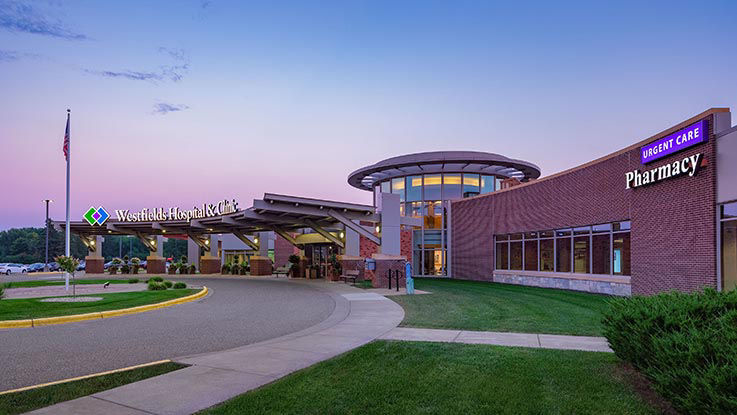 An image of Westfields Hospital & Clinic. It has a wide driveway leading up the main entrance. Signs on the side of the building advertise the urgent care and pharmacy at this location.