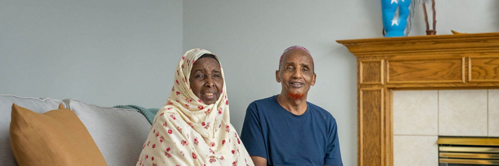 A Somali man and Somali woman smile as they sit on a couch in their home