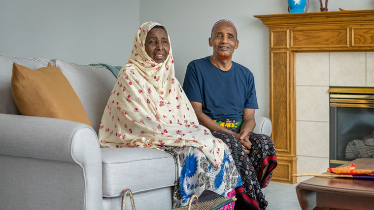 A Somali man and Somali woman smile as they sit on a couch in their home.
