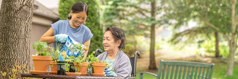 A young Asian woman stands and pours potting soil into a pot on a table outside, while an older Asian woman in a wheelchair smiles and looks on.
