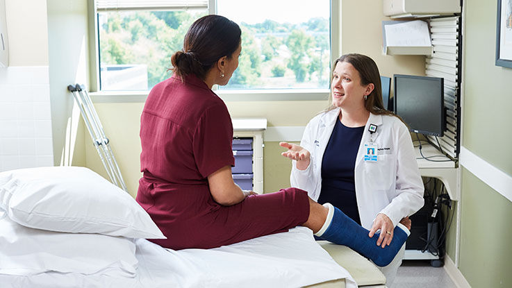 A patient with a cast on her leg sits on the examination table. Her doctor talks with her about treatment recommendations.