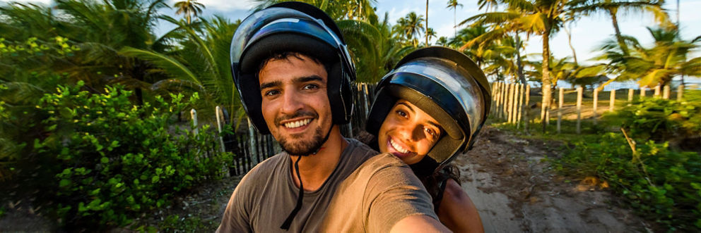 A young man and woman pose for a selfie on a motorbike with palm trees and the ocean in the background.