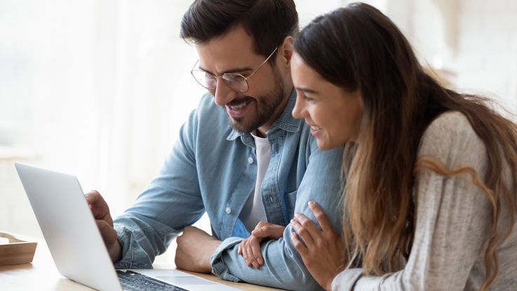 A man and woman sitting together at a table in their home read information from a laptop computer