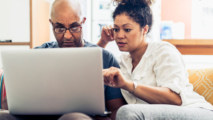 Sitting on their living room couch, a husband and wife research information on their laptop.