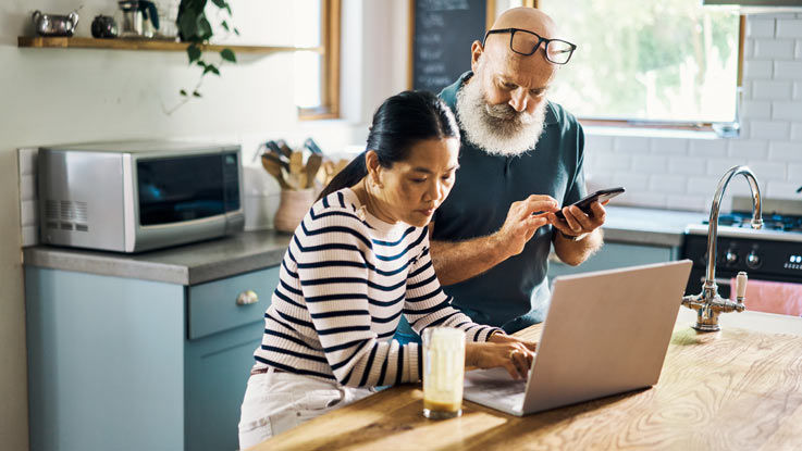 A married couple researches information in their kitchen, the husband on his phone and the wife on her laptop.