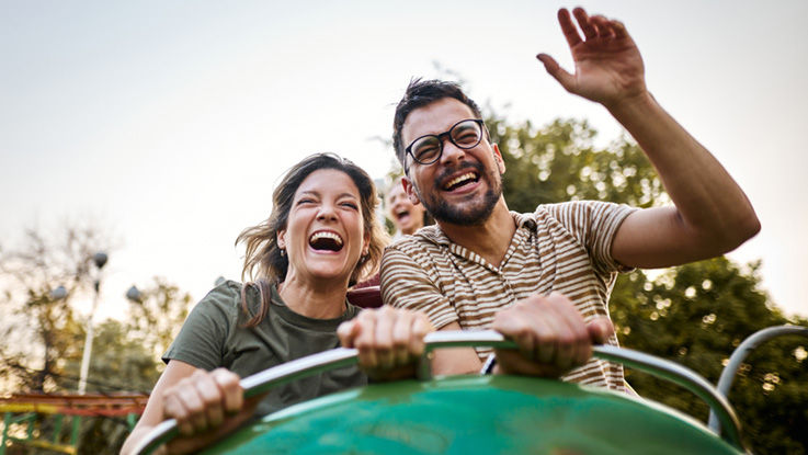 As the sun sets on an amusement park, a young man and women are smiling and laughing as they ride in the front seat of a roller coaster.