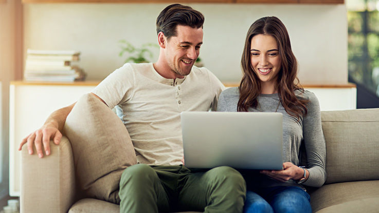 Sitting on their living room couch, a young man and woman smile as they look at their laptop.