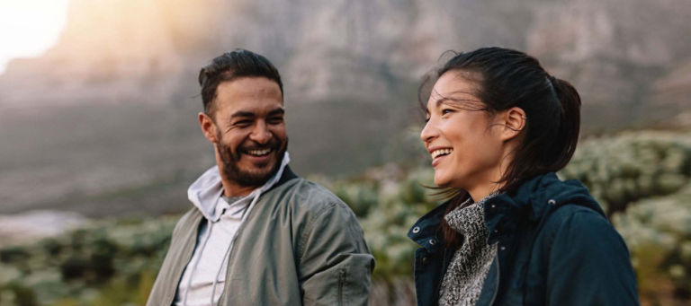 A man and woman hiking outdoors smile and laugh as they talk to each other