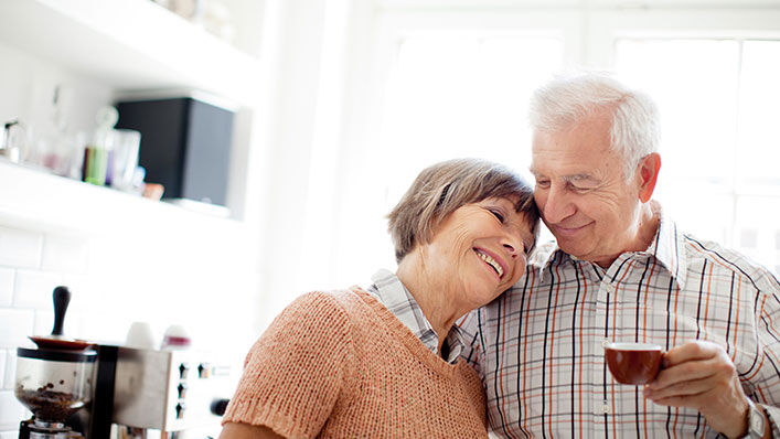 A smiling older woman rests her head on her husband’s shoulder in their kitchen.