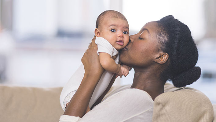 A beautiful young African American mother gently holds her infant daughter up in the air with both hands and kisses her cheek. The baby's eyes are wide open and she looks happy. They are sitting on a couch in their living room.