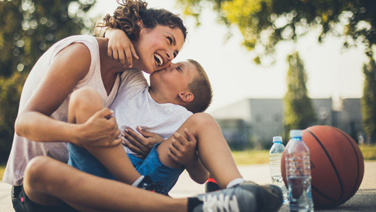 Sitting down on a basketball court and drinking water after a game, a young boy playfully kisses his mother on the cheek while being held in her arms.