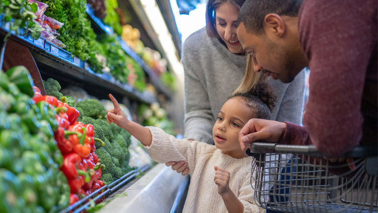 Standing with her parents in the produce aisle of a grocery store, a young girl points to a display of bell peppers.