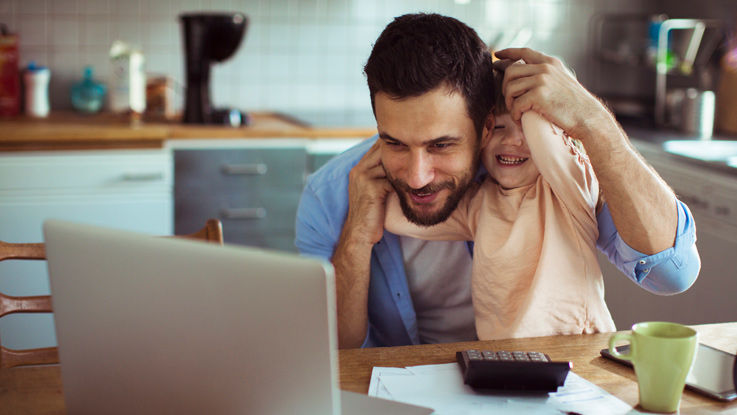  A young father sitting at a kitchen table looks at a laptop. His toddler wraps his arms around the father’s head.