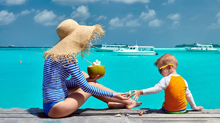 A mother drinks from a coconut while her son plays with seashells as they enjoy a day at the ocean. They're both wearing long-sleeve swimsuits.