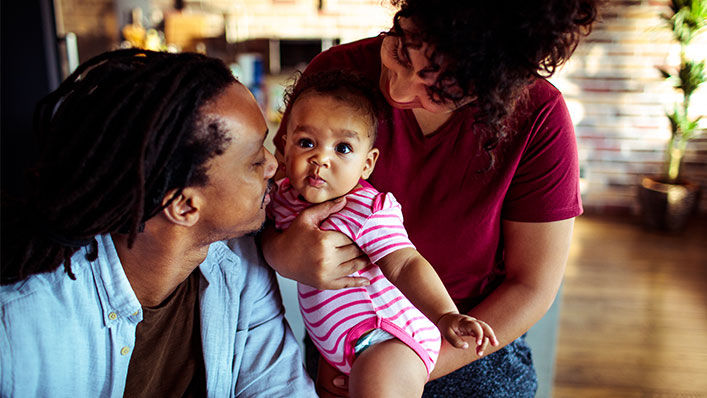 A mom holds her infant daughter while the dad leans in to kiss the baby's cheek.