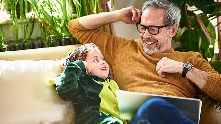 A father and his young daughter sit together on a couch, smiling at each other.