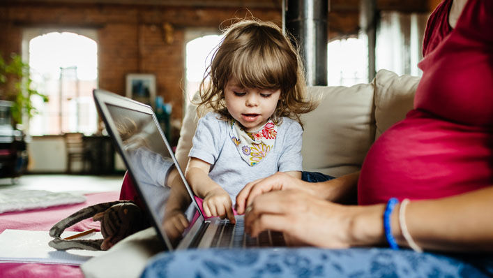 A woman works on her laptop while sitting on a couch in her home as her daughter plays nearby