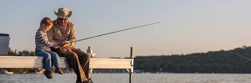 An older man and a boy sit on the side of a dock holding a fishing pole.