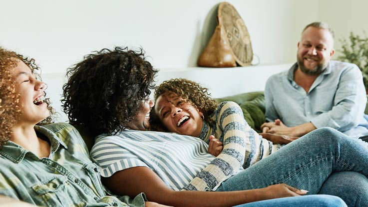 Family of four sit on a couch, hugging and laughing.