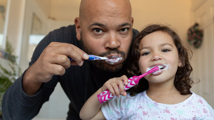 Father and young daughter stand side by side brushing their teeth.