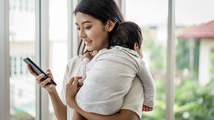 A woman holds a baby while using her other hand to check in on her phone.