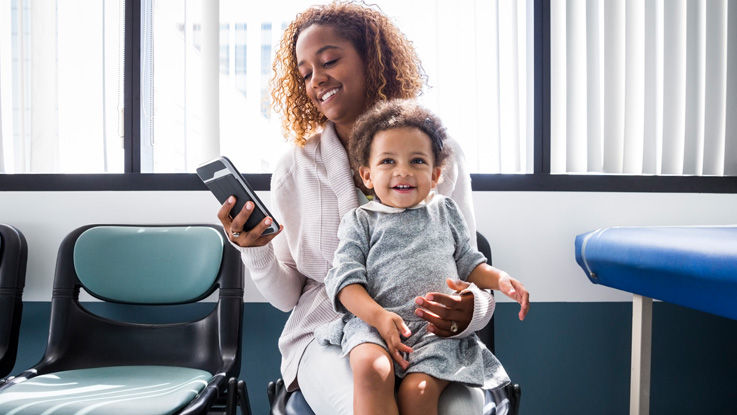 Sitting in a clinic waiting room, a young mother smiles at the phone in her hand while her smiling young daughter sits on her lap.