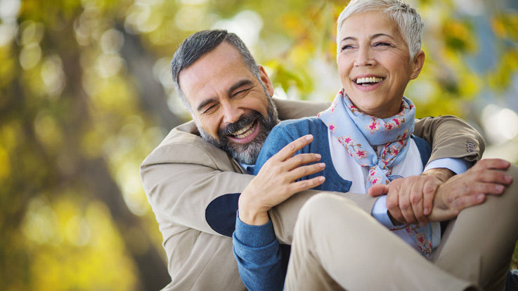 A man laughs as he hugs his smiling wife while they both walk outdoors.