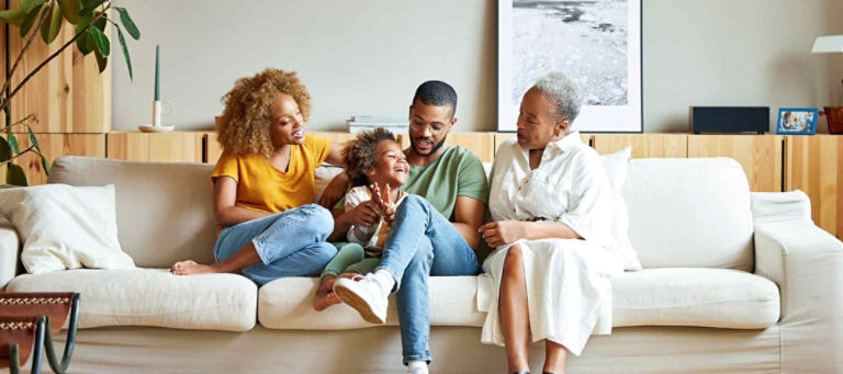 Three generations of a family – grandmother, parents and their young son – sit and share a laugh together on the couch.
