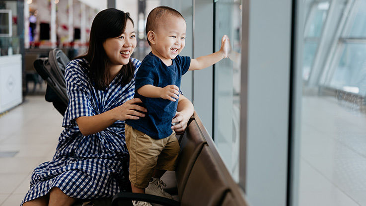 Sitting on an airport bench, a smiling young mother and child look out the window for an airplane.