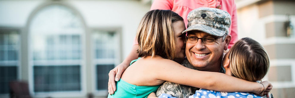 A man in military fatigues returning from overseas deployment smiles as his young daughters hug him