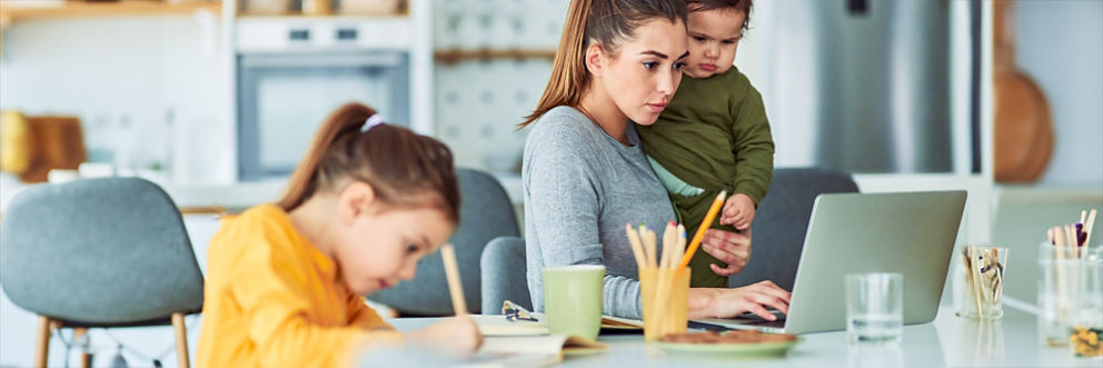 A woman sits at her kitchen table holding a baby while on her laptop. A young girl sits next to her while writing with a pencil.