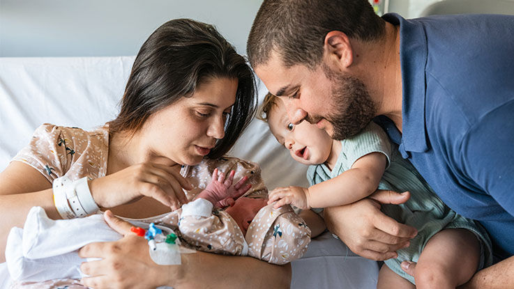 A family together: Dad holds his toddler up to meet the new baby in Mom’s arms.