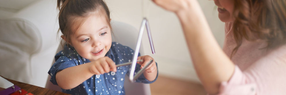 A toddler intently focused on hitting the triangle instrument her mother is holding with a small metal rod.