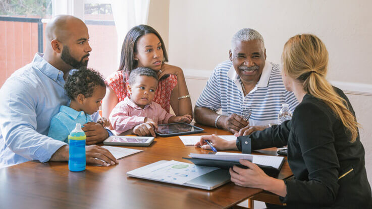 At a table, a husband, wife and two toddlers sit with their smiling grandfather as they talk with a younger woman over paperwork, tablets and a laptop.