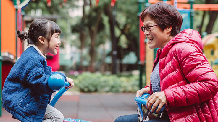 A grandmother and her young granddaughter laugh and smile as they play together on a seesaw at a playground