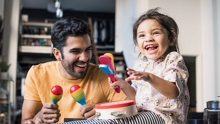 A father and his toddler daughter laughing and playing with various brightly colored musical instruments together.