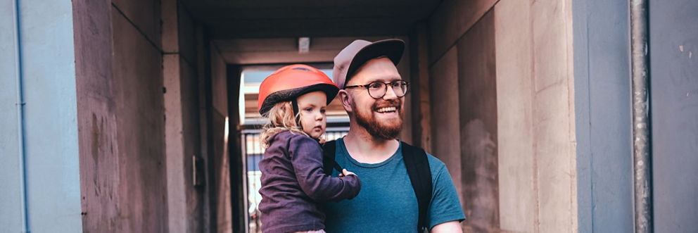 A father carries his toddler daughter who wears a bike helmet.