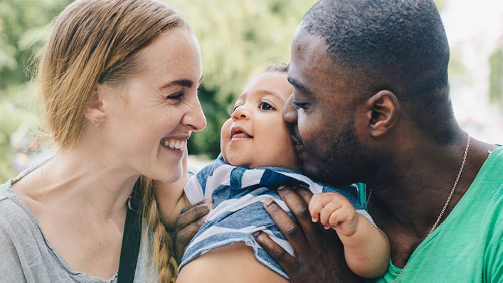 A young woman smiles and laughs as her husband kisses their baby.