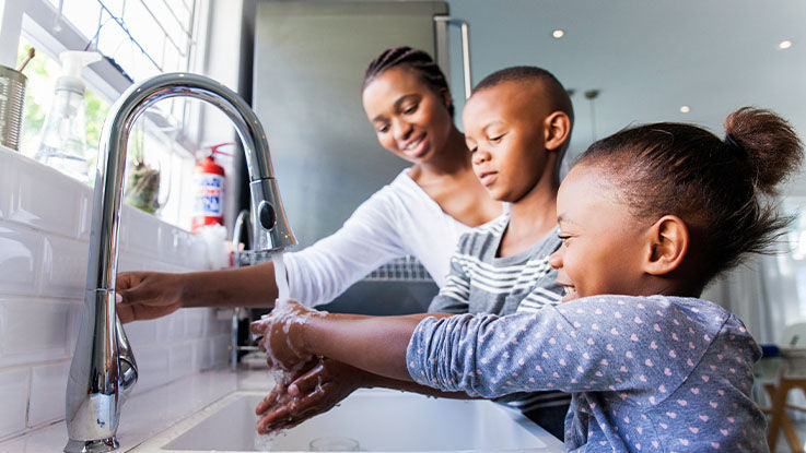 A mom helps her two young children wash their hands at the kitchen sink.