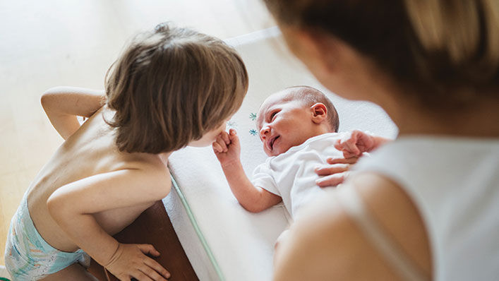 An infant grins and reaches out to their older brother while their mother looks on.