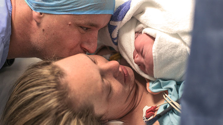 After a C-section, a mother cuddles her newborn on her chest. The baby’s father gazes happily at his baby and kisses his partner on her cheek.