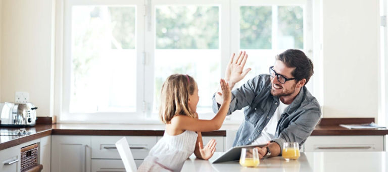 Sitting at their kitchen table during breakfast, a father gives his young daughter a high five while holding a tablet in his other hand.