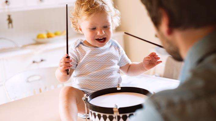 A father and his toddler daughter bang on a toy drum together.