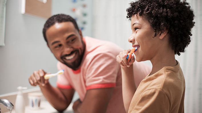 At their bathroom sink, a father is holding his toothbrush, smiling at his son while he is brushing his teeth. 