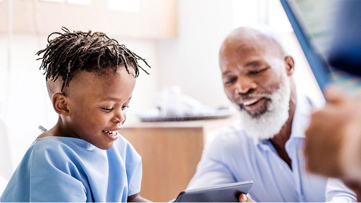 In a hospital room, a boy plays on a tablet while his father looks on.